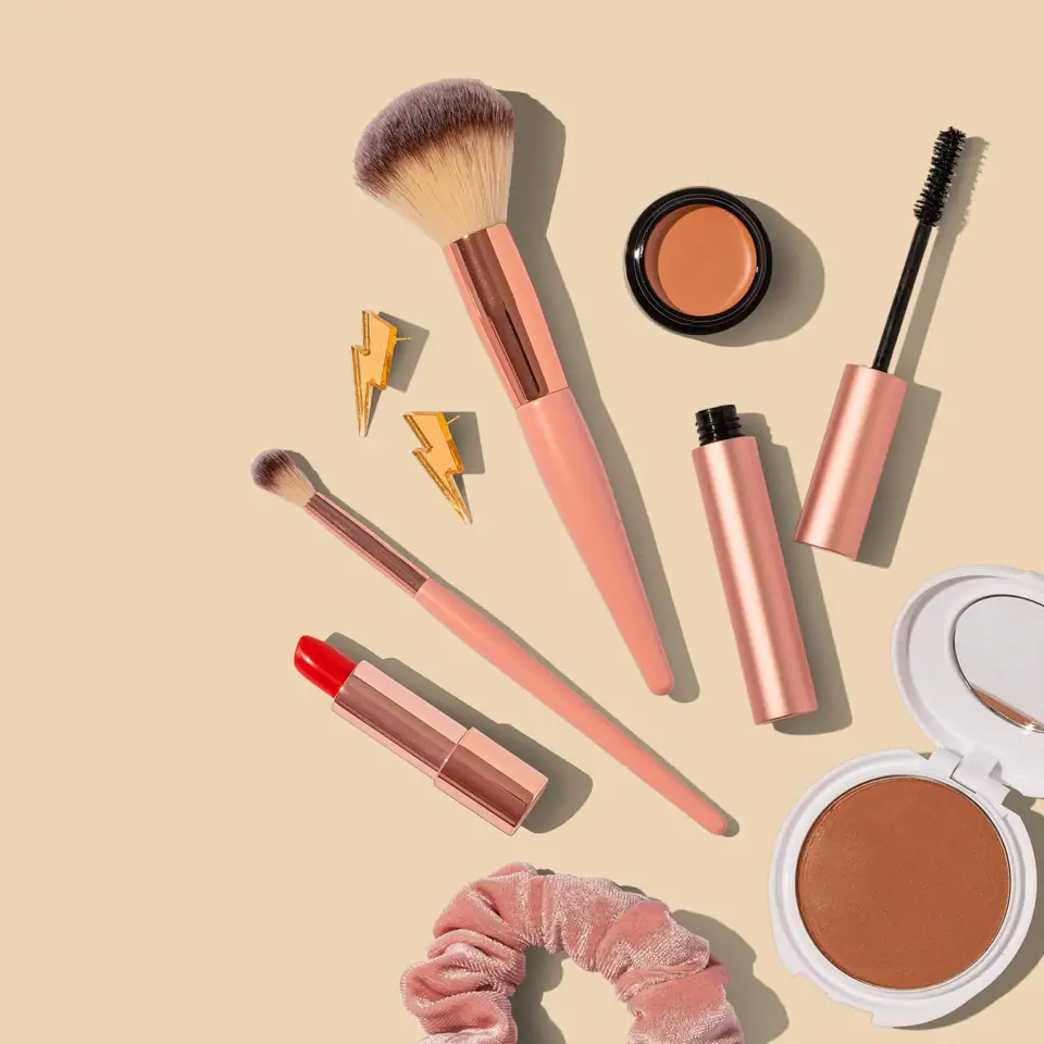 A flat lay of makeup brushes, a lipstick, mascara, blush and a compact on a peach background